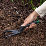 Hand holding an Expert Gardener poly cultivator digging into loose soil and mulch in a garden bed.