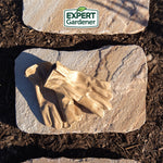 Pair of tan Expert Gardener leather gloves resting on a flat stone with the brand logo above, photographed outdoors on mulch.