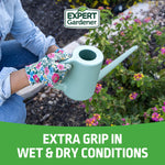 Lifestyle image of person wearing Expert Gardener floral gloves holding a mint-green watering can near flowering plants