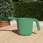 Green ribbed resin watering can placed on a wooden surface outdoors with plants and hedges in the background.