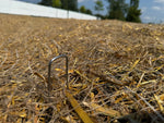 A U-shaped metal landscape pin partially driven into straw-covered ground, demonstrating outdoor use.