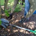 Shovel lying on soil while a person kneels to plant greenery, showing shovel handle and blade in use