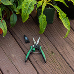 Garden snips placed on wooden deck beside potted plants with protective blade cap nearby