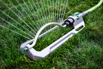 Oscillating lawn sprinkler in use on grass with water spraying from the arched metal spray bar.