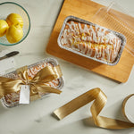 Overhead styled photo of two baked and iced loaves in disposable aluminum pans with clear lids and decorative ribbon