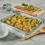 Filled foil casserole pan on a marble counter with serving spoon, cutting board and parsley garnish
