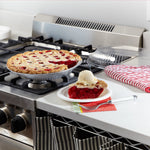 Kitchen scene showing a baked cherry pie in an EZ Foil pan on a stovetop with a plated slice and clear lid