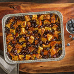Top-down view of a foil pan filled with bread and blueberries placed on a wooden board and cloth napkin