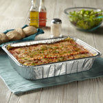 Baked lasagna in an aluminum disposable pan on a placemat with bread and salad in the background
