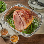 Disposable aluminum roaster pan holding a glazed ham on bed of green beans with serving dishes nearby.