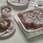 Square aluminum foil pan holding a sliced chocolate cake with a plated slice, fork and bowl of chocolate nearby