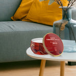Red Febreze tin candle on a round side table in a living room setting beside a glass vase