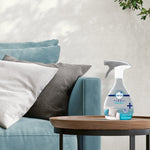 Febreze Fabric Heavy Duty spray bottle placed on a round wooden side table beside a teal couch with pillows.