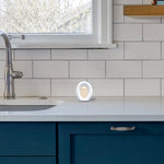 Febreze oval air freshener device placed on a kitchen countertop near a sink in a lifestyle setting