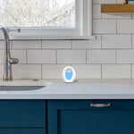 Febreze Small Spaces device placed on a kitchen countertop near a sink and subway tile backsplash in a lifestyle shot