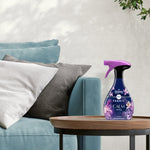 Febreze Fabric Calm spray bottle placed on a round wooden side table next to sofa cushions in a neutral living room scene