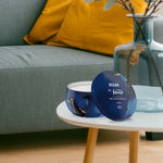 Styled room scene with a Febreze Ocean tin candle on a white side table in front of a sofa and decorative glass vase.