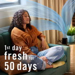 Person relaxing on a sofa while a Febreze PLUG scent booster operates nearby with stylized scent waves and freshness claim