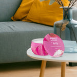 Febreze Romance & Desire pink candle tin displayed on a small round table in a living room setting with sofa and decorative vase