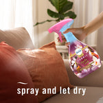 Hand holding a Febreze Vanilla Suede fabric spray above a textured throw pillow in a sunlit living room.