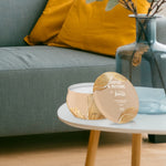Staged home scene with beige Febreze Soothe & Restore tin candle on a small white table beside a sofa and glass vase