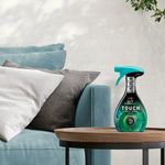 Product-in-context photo of a Febreze Unstopables Touch bottle on a wooden side table beside sofa cushions