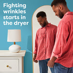 Man inspecting a red shirt in front of a mirror beside a lamp and dresser, illustrating wrinkle reduction from the dryer.