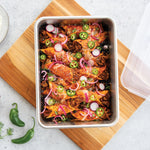 Aluminum baking pan filled with seasoned food and sliced jalapeños, shown on a wooden board with a translucent lid to the side