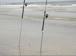 Two fishing rods set in sand with metal or PVC sand spikes supporting the rods on a beach with waves in background