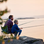 Father and child fishing at a lakeside with a collapsed gray collapsible basin placed on the rock beside them.