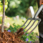 Close-up lifestyle scene of a Fiskars four-tine pitchfork lifting mulch in a garden bed with blurred greenery background.