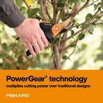 Hand holding Fiskars PowerGear2 pruner cutting a small woody stem with garden background and PowerGear text