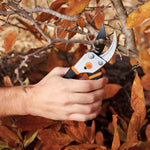 Hand holding a Fiskars bypass pruner trimming a branch amid autumn leaves in an outdoor scene