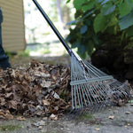 Close-up of Fiskars rake head and metal tines pulling dried leaves near garden foliage.