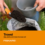 Hand using a Fiskars orange-and-black trowel scooping potting soil from a bag over a garden scene with orange product panel