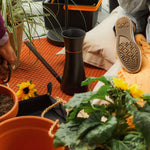 Lifestyle photo showing Fiskars watering can among pots and gardening tools on an indoor rug near plants