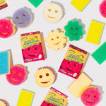 Flat lay of assorted Scrub Mommy smiley sponges and two boxed Scrub Mommy products on a white background