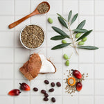 Flat lay of natural ingredients including coconut pieces, seeds, olives and palm fruits on tiled surface.