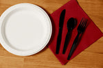 Top-down view of a paper plate next to a red napkin with black disposable knife, spoon and fork arranged on it