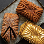 Two fluted loaves and a gold-colored loaf pan displayed on a decorative baking sheet, one loaf dusted with powdered sugar