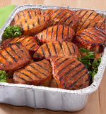 Close-up of a foil roaster pan filled with grilled, glazed meat pieces and garnishes on a wooden surface