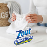 Folded white shirt being handled on a bed with large Zout logo and 'Stains Gone. Guaranteed!' branding overlay