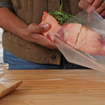 Hands placing a large raw cut of meat and rosemary into a FoodSaver textured vacuum bag over a wooden countertop.