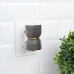 Ribbed dark gray fragrance plug inserted in a wall outlet beside a small green plant on a white tile wall