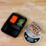 Open black three-compartment meal prep container with clear lid and a kitchen scale showing skewered meat on a wood surface