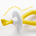 Macro view of the yellow Frida Baby training toothbrush resting on its white removable safety stopper with headline text