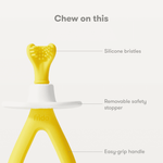 Close-up product callout of a yellow Frida Baby training toothbrush showing silicone bristles, safety stopper, and easy-grip handle