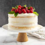 Frosted layered cake topped with strawberries, raspberries and mint on a white pedestal against a dark backdrop