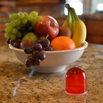 Red translucent fruit fly trap on a kitchen countertop in front of a bowl of assorted fruit