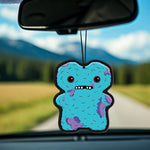 Blue Fuggler monster air freshener with red eyes and purple patches hanging from a rearview mirror against a blurred road and field backdrop.